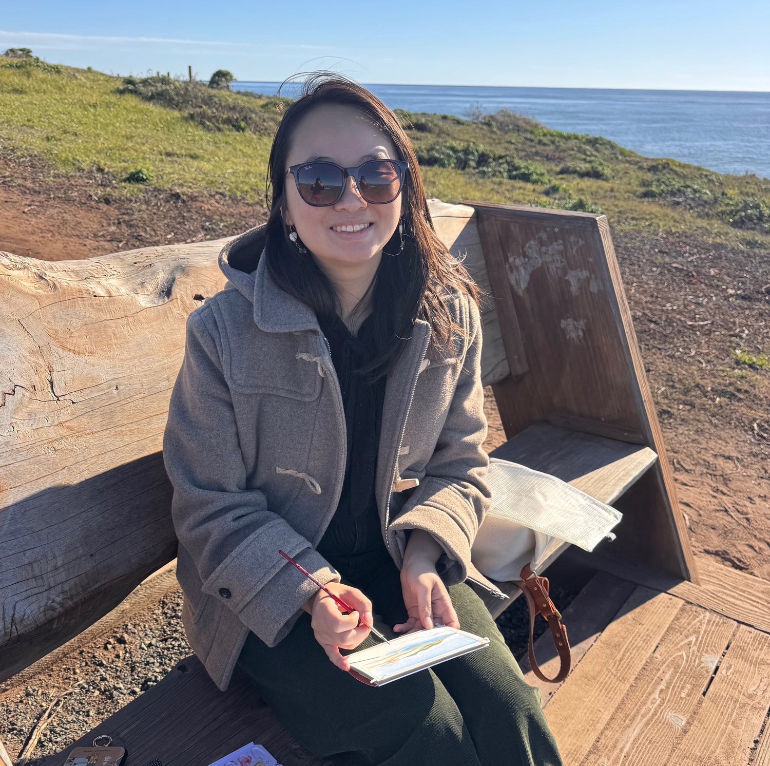 Woman sitting on a wooden bench by the ocean, holding a small painting.