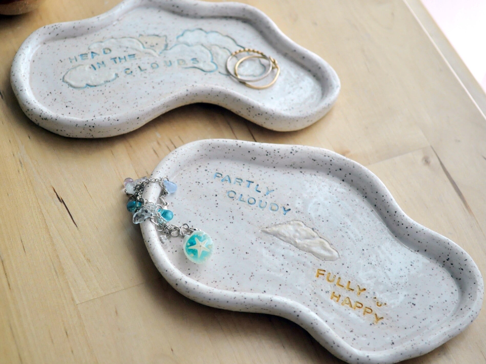 Two cloud shaped ceramic trinket dishes on a wooden surface with jewelry.