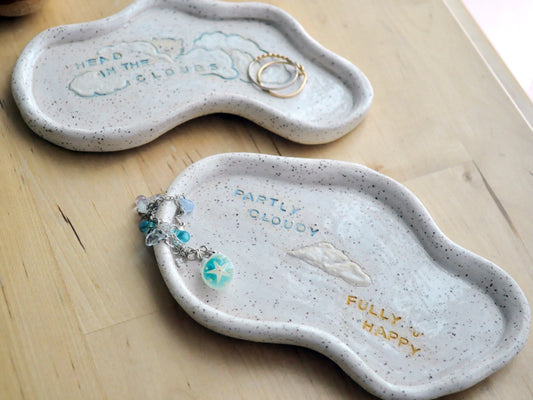 Two cloud shaped ceramic trinket dishes on a wooden surface with jewelry.