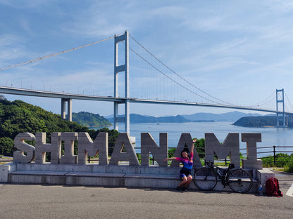 Woman posing in front of a large 'SHIMANAMI' sign with her bike, with a bridge and scenic background on the Shimanami Kaido cycling route in Japan.